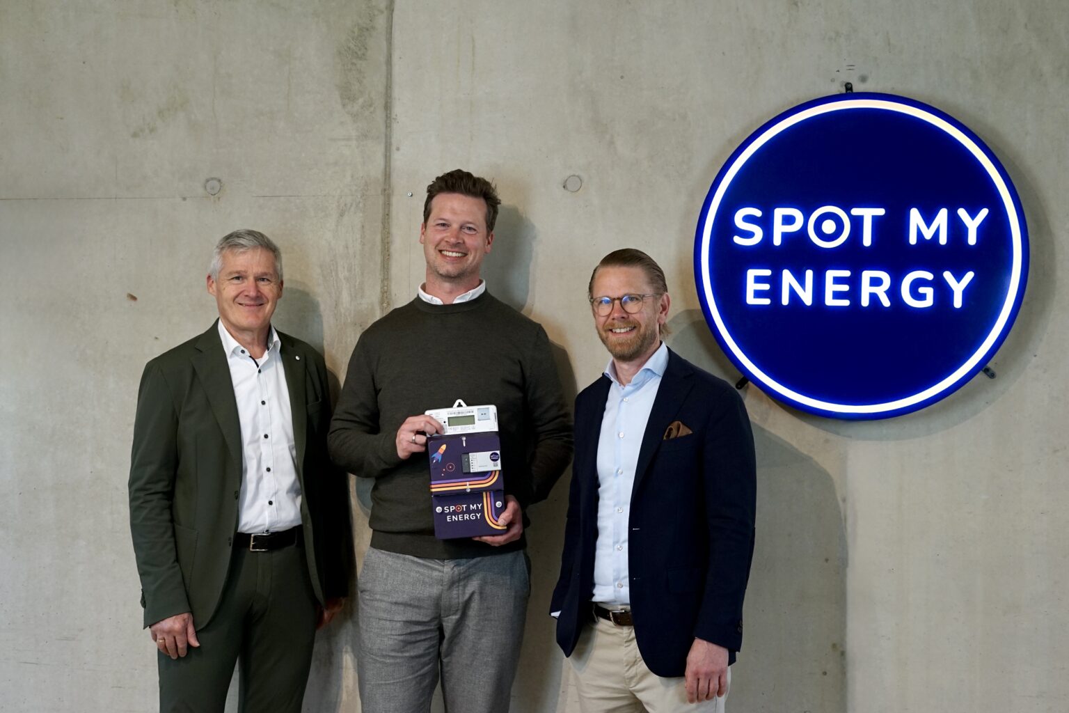 Three men in business attire stand by a concrete wall beside a blue Spot My Energy sign; center man holds a product box.