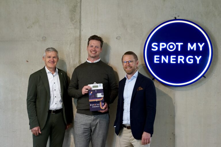 Three men in business attire stand by a concrete wall beside a blue Spot My Energy sign; center man holds a product box.
